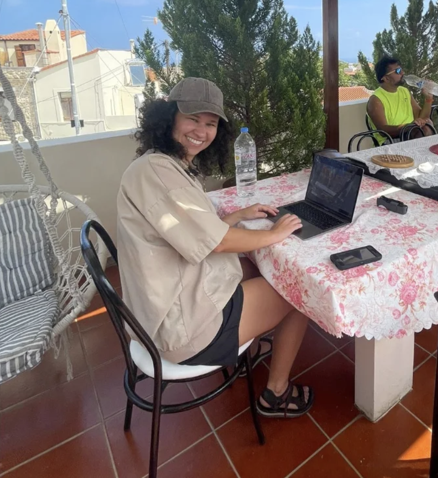 Emma working remotely on her laptop at an outdoor terrace in Greece, wearing a beige shirt and baseball cap, smiling at the camera with Mediterranean buildings in the background